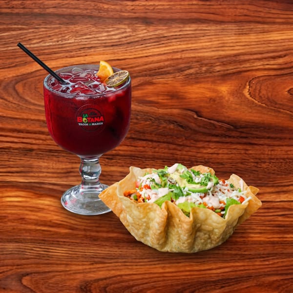 A taco salad in a crispy shell and a red drink with lime and orange on a wooden table.