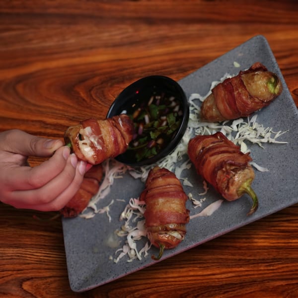 A hand picks up a bacon-wrapped jalapeño popper from a grey plate with dipping sauce.