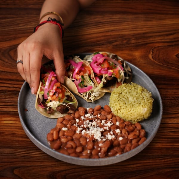A hand reaching for a taco on a plate with two other tacos, beans, and rice.