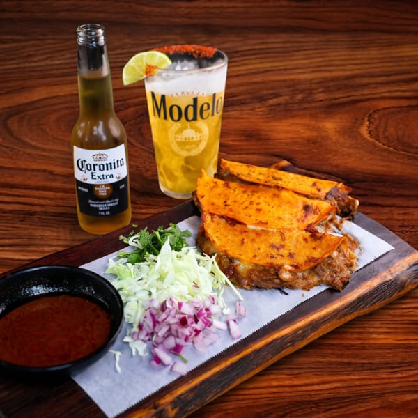 Birria tacos with cilantro, onions, dipping sauce, a Modelo beer, and a Coronita on a wooden board.